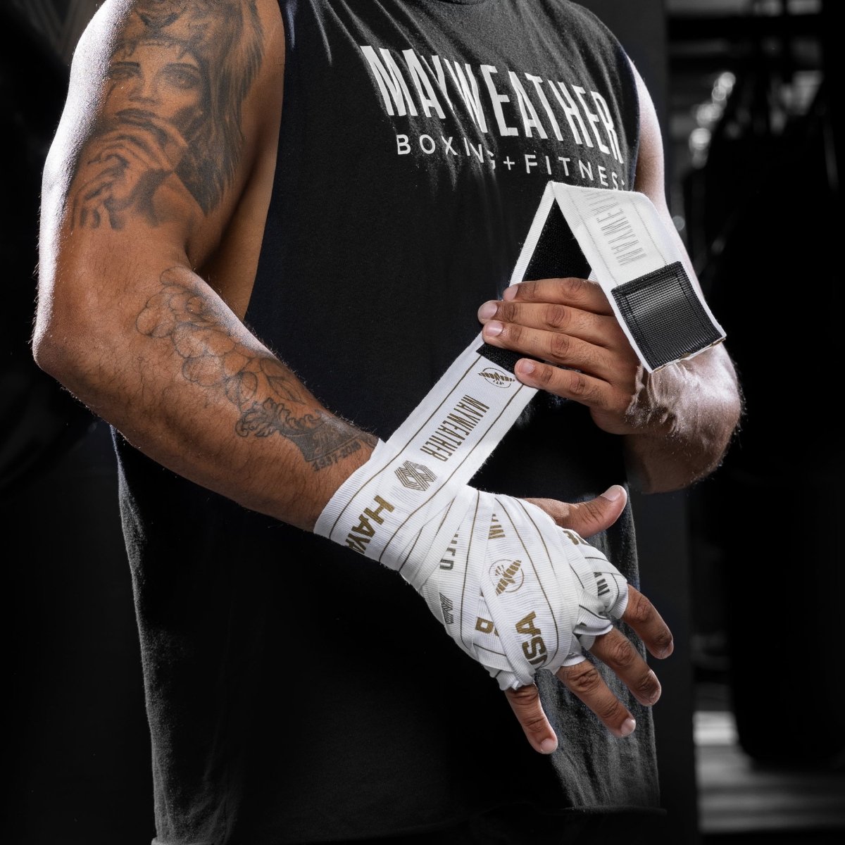 Vista frontal da mão de um lutador com uma bandagem completa usando a bandagem elástica Mayweather Hayabusa de 180 polegadas branca e dourada, com o logotipo 'MAYWEATHER' visível na área do pulso